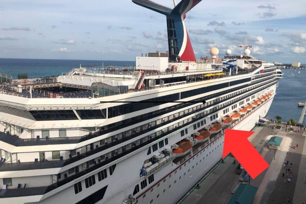 This image shows a Carnival cruise ship docked at a port, with a red arrow pointing to a specific row of balcony cabins. The ship features Carnival's signature red and blue funnel, lifeboats along the lower deck, and various amenities visible on the upper deck. The photo highlights the ship's design and accommodations.