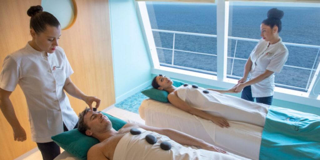 A couple enjoying a relaxing spa treatment with hot stones on a Carnival cruise ship, with therapists providing skilled services, all set against a serene ocean backdrop visible through a large window.