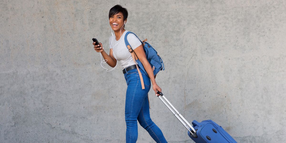 A cheerful young woman in casual attire listens to music on her phone while walking with a rolling suitcase, exemplifying the ease of modern travel.