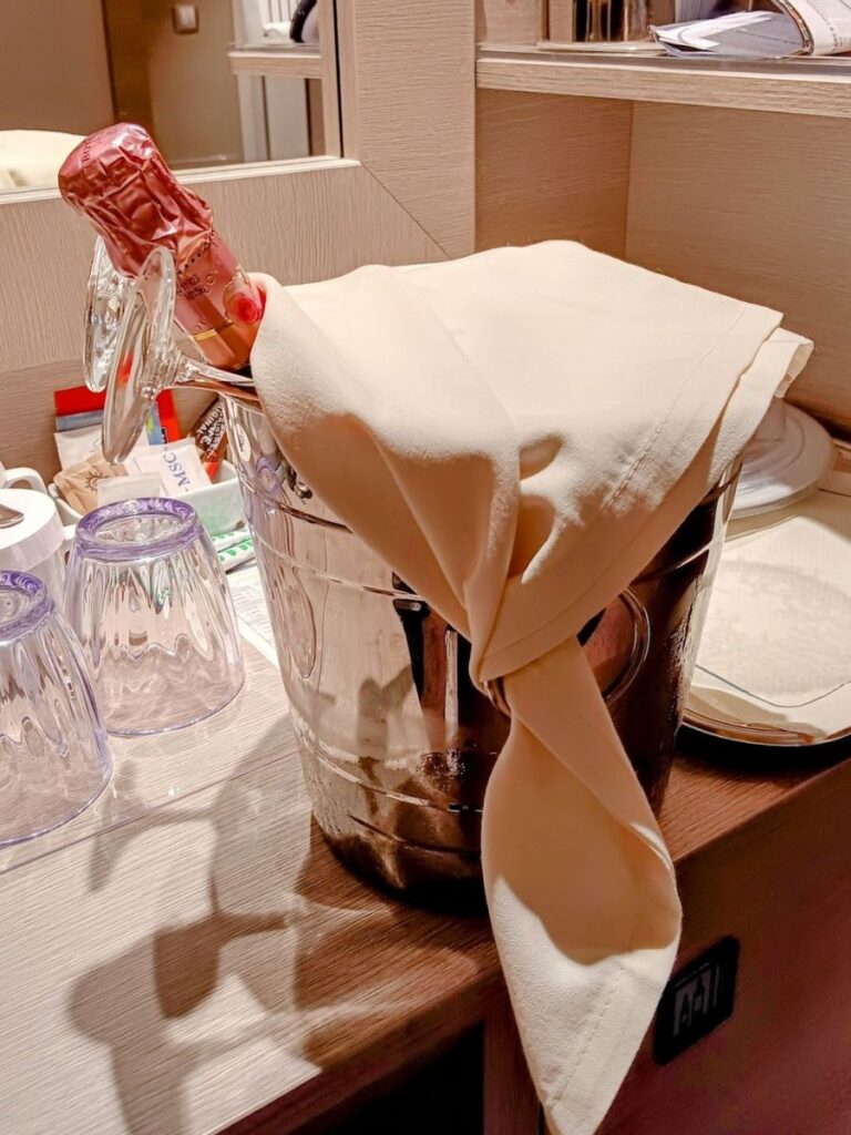 An intimate celebration setting featuring a chilled champagne bottle wrapped in a beige linen napkin, peeking out of a shiny ice bucket, with clear crystal glasses nearby, all resting on a wooden surface, hinting at a romantic or celebratory occasion in an upscale ambiance.