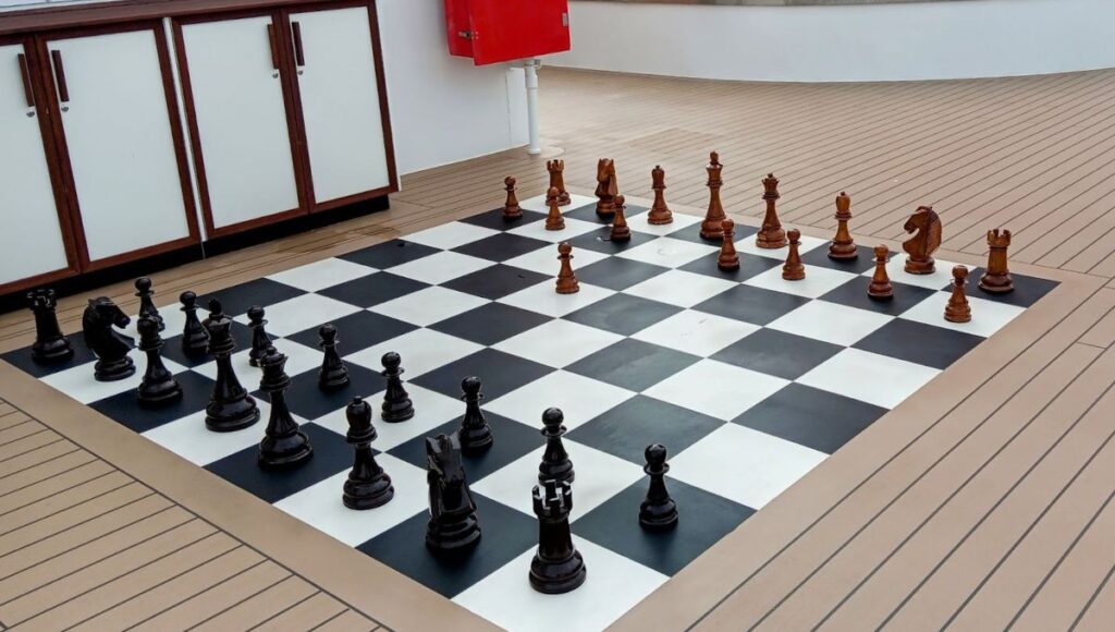 The image shows a life-sized chessboard on Cunard's Queen Anne cruise ship. The large black and white squares form the playing surface, with oversized wooden chess pieces set up for a game. The chess pieces are carved in black and brown, contrasting with the ship's light-colored deck. The setting offers a fun and interactive activity for passengers, blending leisure with a classic game in a spacious outdoor or semi-outdoor area.