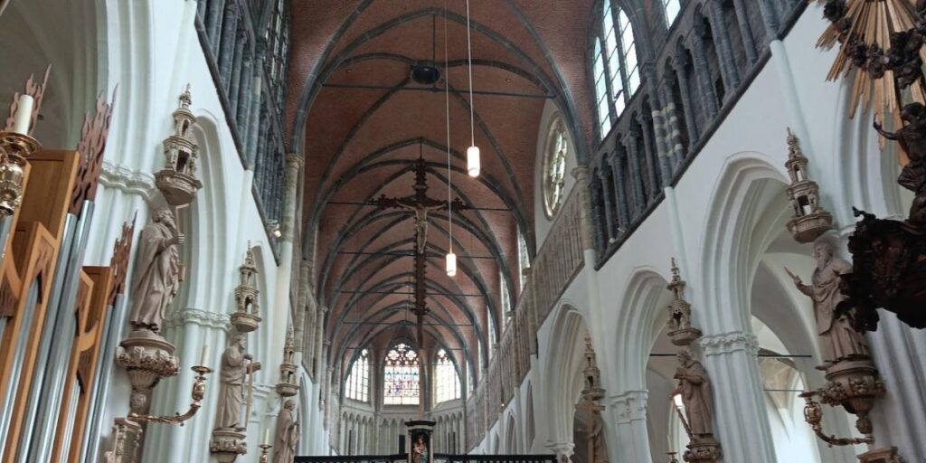 The interior of a grand cathedral, featuring high vaulted ceilings, ornate statues, and intricate architectural details. The nave is lined with tall columns and decorative elements, leading to a stained glass window at the far end, creating a majestic and reverent atmosphere.