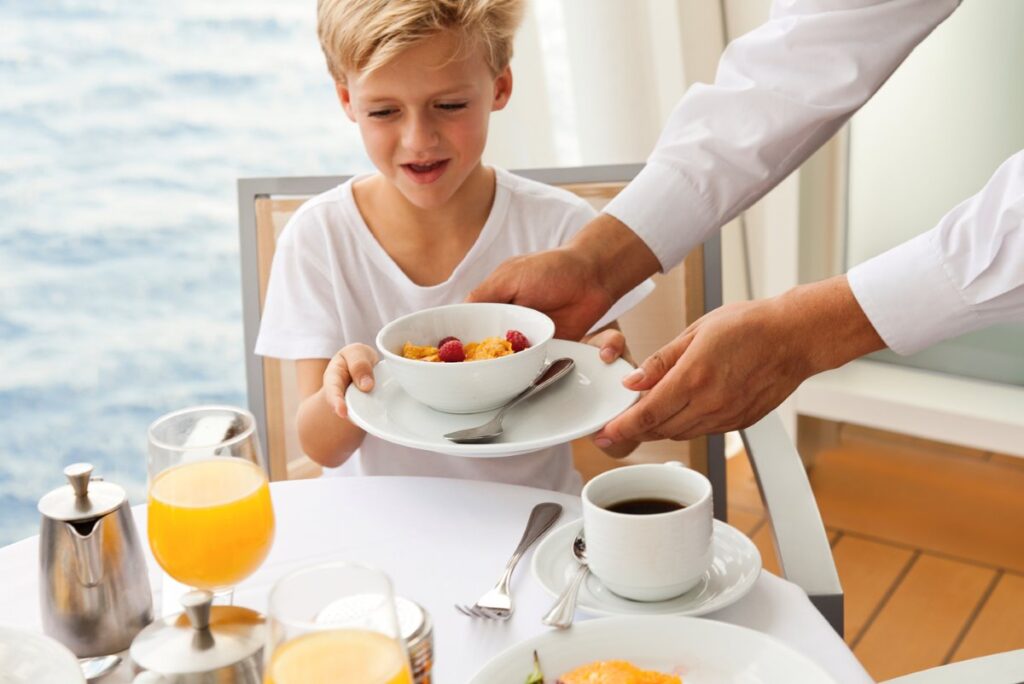 A delighted young child about to receive a bowl of fresh cereal topped with raspberries in a Royal Caribbean suite. The breakfast table is neatly set with a glass of orange juice, a cup of coffee, and an inviting ocean view in the background, encapsulating a luxurious family dining experience at sea.