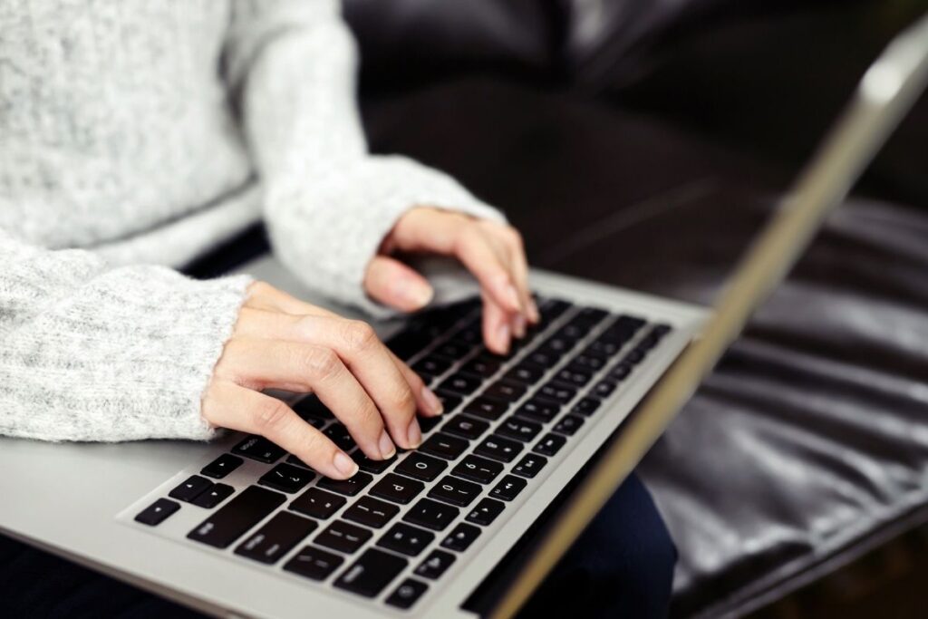 This image shows a close-up view of hands typing on a laptop keyboard. The person is wearing a cozy, light-gray sweater, suggesting a casual or relaxed setting. The laptop's sleek design and focused activity capture the essence of modern work or leisure in a digital environment.
