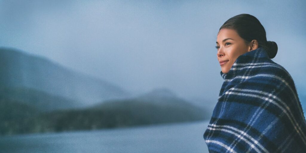 A woman wrapped in a cozy plaid blanket stands against a misty mountainous backdrop, suggesting a chilly outdoor setting.