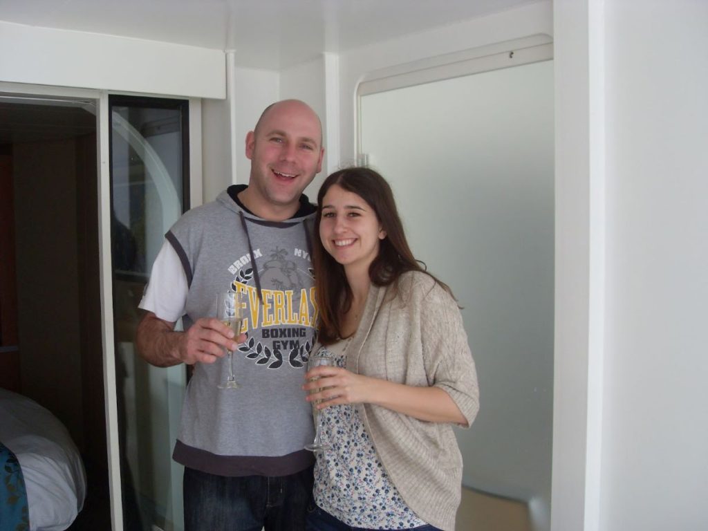 A cheerful couple holding champagne flutes in a Royal Caribbean stateroom, standing by the balcony door, embodying a relaxed cruise atmosphere.