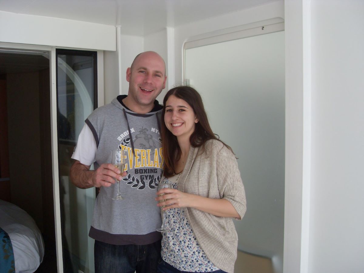A cheerful couple holding champagne flutes in a Royal Caribbean stateroom, standing by the balcony door, embodying a relaxed cruise atmosphere.