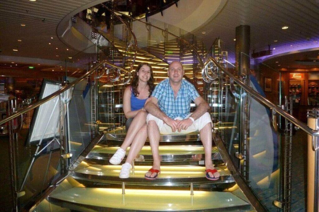 A relaxed couple sits on the golden-lit steps inside the 'Oasis of the Seas' cruise ship, surrounded by the gleaming chrome and glass of the ship's modern interior, enjoying their casual vacation time.