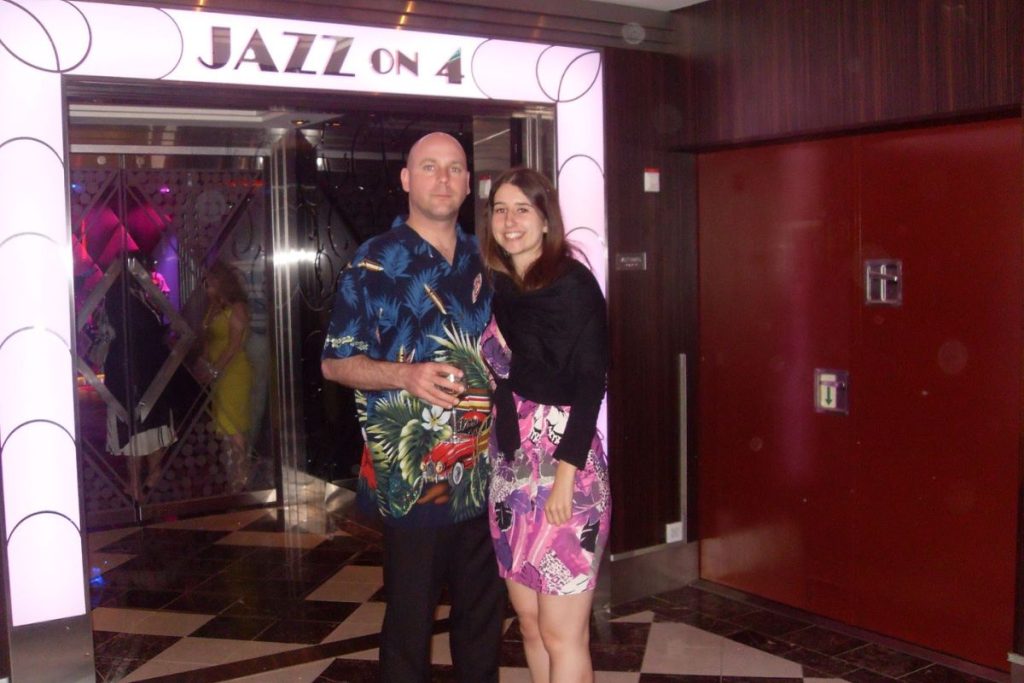 A couple stands outside the 'Jazz on 4' club on a Royal Caribbean cruise, ready for a theme night, the man in a tropical print shirt and the woman in a patterned dress, both smiling and anticipating an evening of music and entertainment.