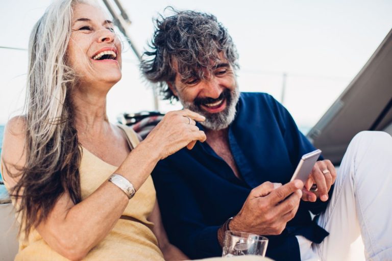 An elderly couple joyfully using a smartphone together on a cruise ship, with the woman laughing and the man smiling broadly, both casually dressed and enjoying their time at sea.
