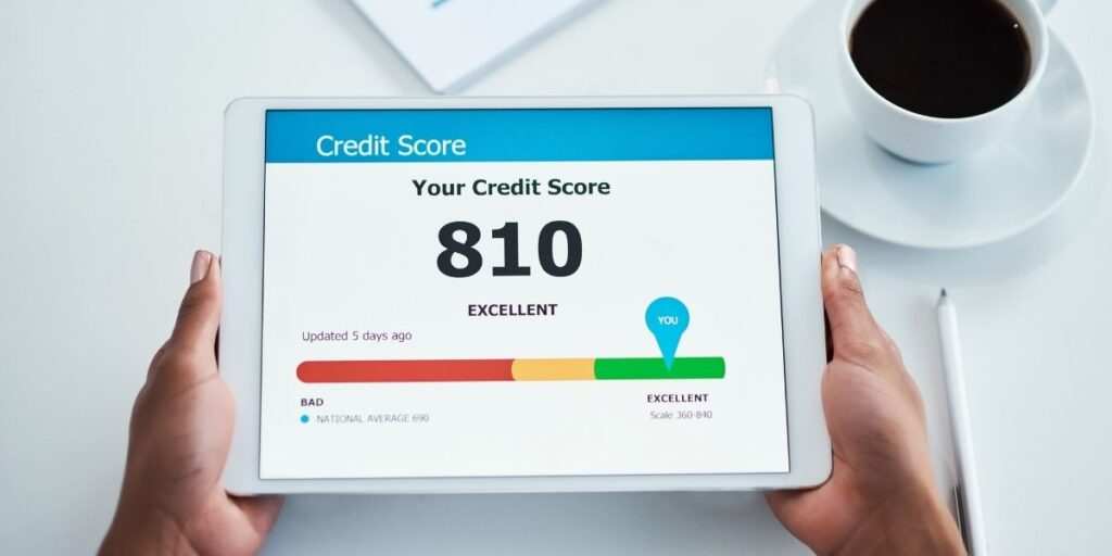 Person holding a tablet displaying a credit score of 810, labeled as "Excellent" on a color scale ranging from red (bad) to green (excellent), with a coffee cup and pen visible on the white table in the background.