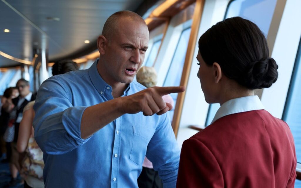 An intense interaction on a cruise ship, where a bald man in a blue shirt sternly points his finger at a female staff member dressed in a red and white uniform. The scene takes place beside large windows overlooking the ocean, with other passengers visible in the background.