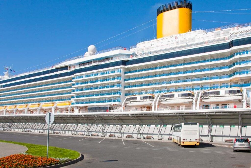 A large cruise ship docked at a port, with a bright yellow funnel and multiple decks visible. In the foreground, a white bus is parked near the terminal, ready for passenger transport. The scene is set under a clear blue sky, indicating a sunny day.