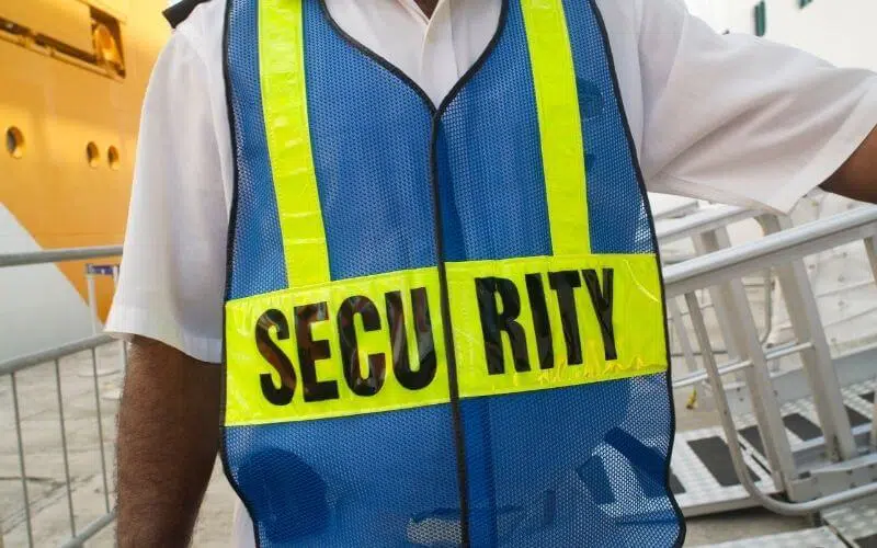 A security guard in a white uniform shirt wearing a blue safety vest with yellow reflective stripes. The vest has the word "SECURITY" printed on it, but the middle letters are missing or obscured, reading "SECU RITY." A cruise ship and metal gangway are visible in the background, suggesting a port or boarding area.