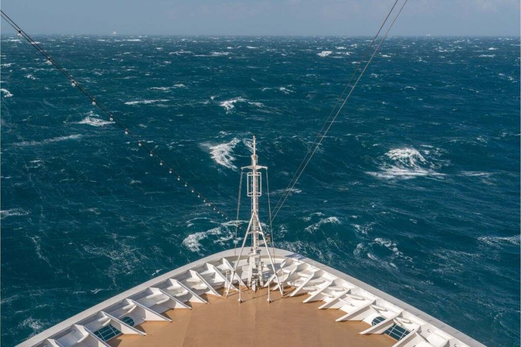 The image shows the bow of a cruise ship as it navigates through rough seas. The ocean is choppy, with white-capped waves visible across the deep blue waters. The ship's deck is seen from above, with the mast and rigging for lights extending over the bow. The contrast between the structured, clean lines of the ship and the wild, turbulent ocean highlights the dynamic nature of cruising through challenging weather conditions.