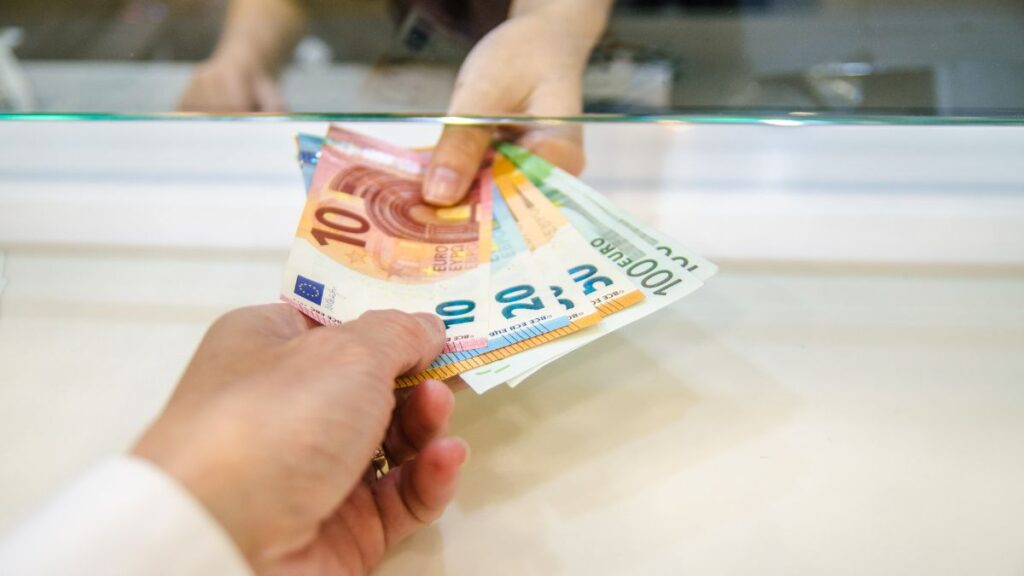 A close-up shot of a transaction at a currency exchange counter, where one person hands over a stack of euro banknotes, including 10, 20, and 100 euro bills, to another person behind a glass partition. The exchange takes place through a small opening in the glass, with the hands of both individuals being the primary focus of the image. The setting suggests a bank, exchange office, or financial service location.
