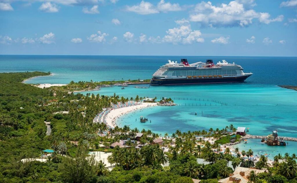 The Disney Wish cruise ship docked at Castaway Cay, Disney's private island, with a bustling beachfront, turquoise waters, and lush greenery under a blue sky.
