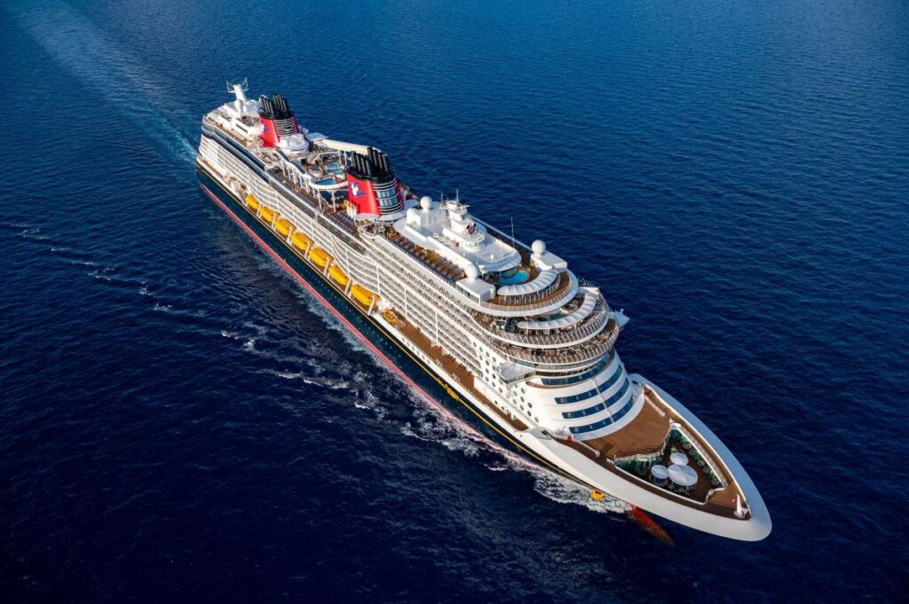 Aerial view of the Disney Wish cruise ship sailing on a calm blue ocean, featuring its distinctive red, black, and yellow color scheme and Disney-themed design elements.