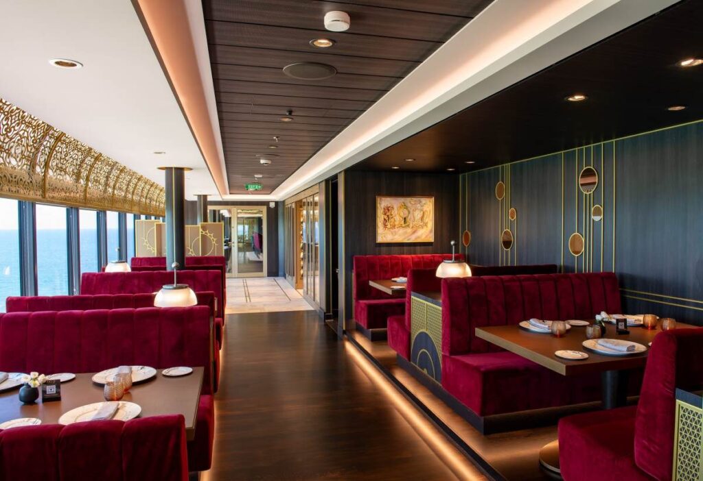 Inside the Palo Steakhouse on the Disney Wish, showcasing elegant red velvet seating, stylish gold and dark wood decor, and a panoramic ocean view from the large windows.