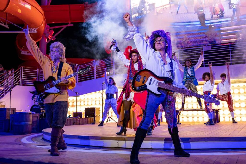 Energetic performers dressed as pirates rocking out with electric guitars at the Pirate’s Rockin’ Parlay Party on the Disney Wish, with dynamic lighting and theatrical smoke adding to the vibrant atmosphere on deck.
