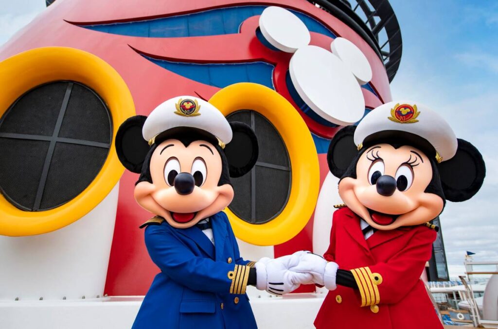 Mickey and Minnie Mouse characters in captain's attire, shaking hands in front of the iconic red, black, and yellow funnel of the Disney Wonder cruise ship, under a cloudy sky.