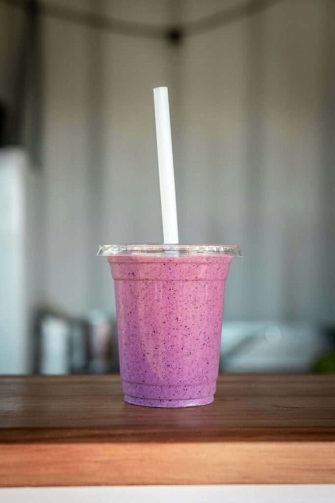 Purple berry smoothie in a clear plastic cup with a lid and straw, sitting on a wooden table with a softly blurred background.