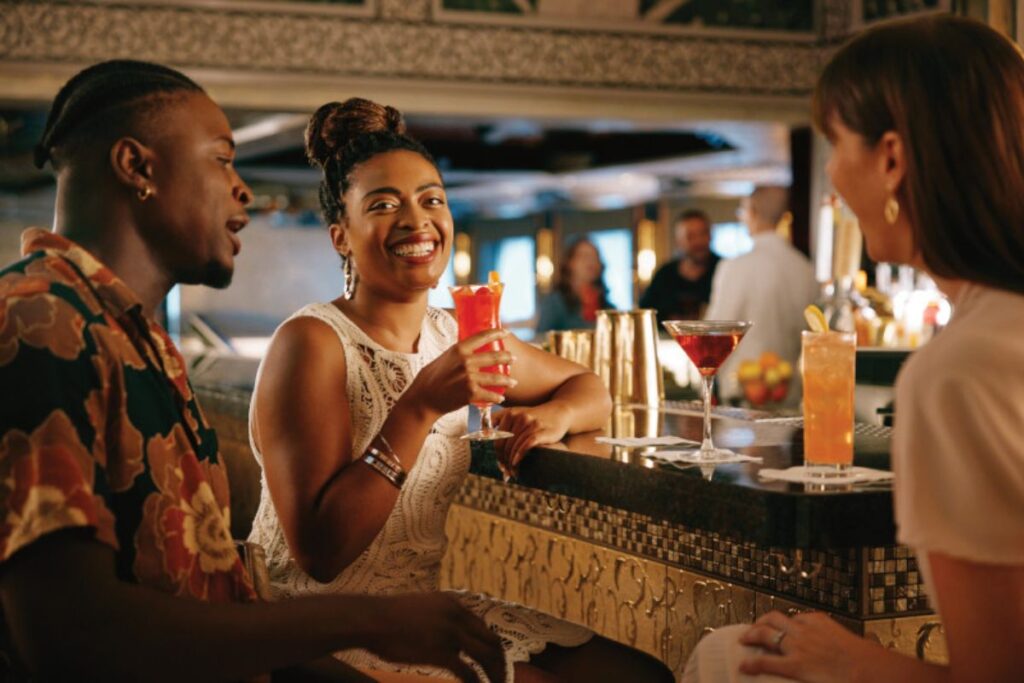 A group of three friends enjoying drinks at a bar, with a woman in the center holding a vibrant red cocktail and smiling at her companions.