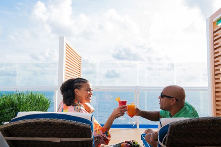 A couple toasting with orange drinks on a cruise ship balcony, enjoying the expansive ocean view under a bright blue sky.