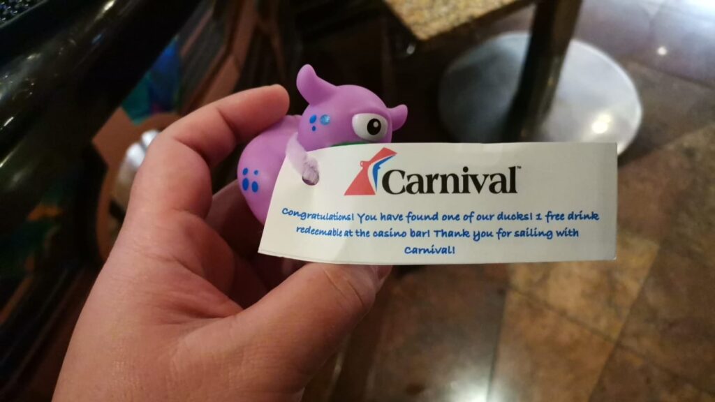 A person holds a small purple rubber duck with blue spots. Attached to it is a tag with the Carnival Cruise Line logo that reads: "Congratulations! You have found one of our ducks! 1 free drink redeemable at the casino bar! Thank you for sailing with Carnival!"