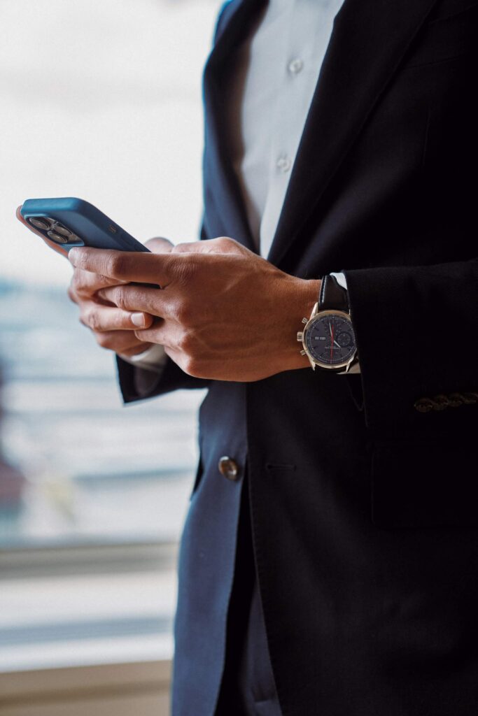 Close-up of a person in a suit holding a smartphone, with a wristwatch visible, against a softly blurred background.