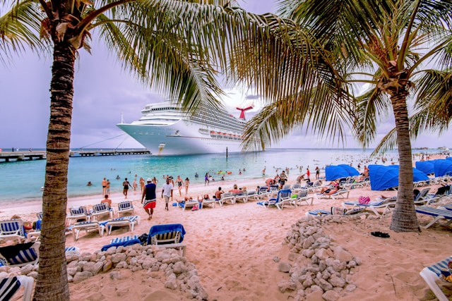Cruise ship docked offshore by a tropical beach with palm trees, lounge chairs, and vacationers swimming and relaxing on the sand.