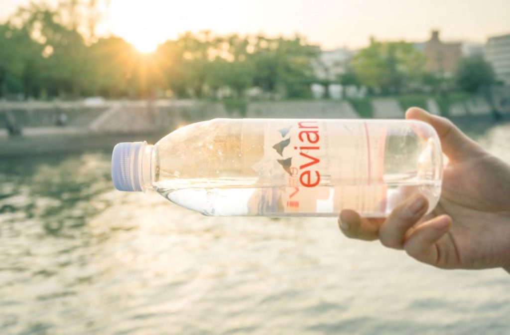 A hand holding a transparent Evian water bottle, with the sun setting in the background creating a serene backdrop over a calm body of water. The scene captures a moment of refreshment in a peaceful outdoor setting.