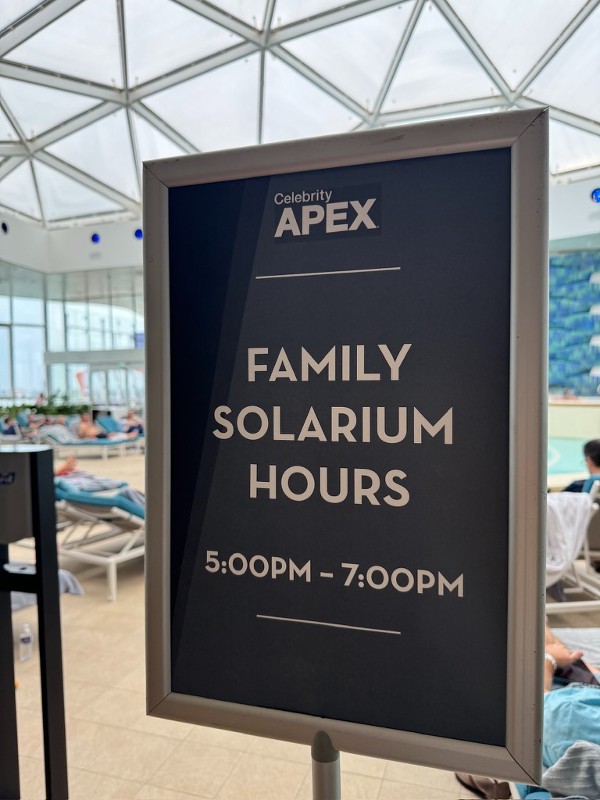 Sign inside the Celebrity Apex cruise ship reads "Family Solarium Hours 5:00PM – 7:00PM," positioned near a pool area with guests lounging on blue cushioned chairs beneath a large glass dome.