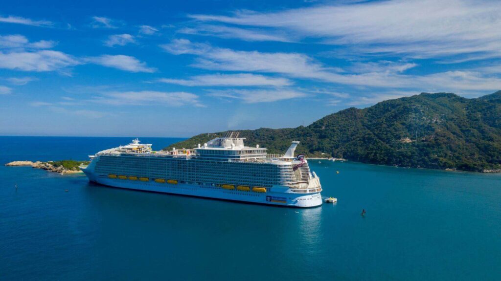 Aerial view of Royal Caribbean's Symphony of the Seas cruise ship sailing in calm blue water near a lush, mountainous island coastline in Haiti.
