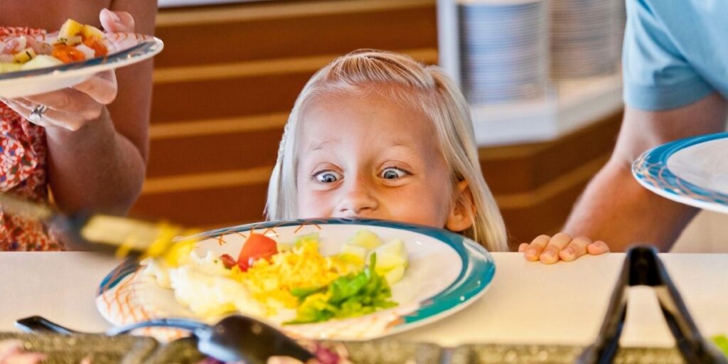 A young child with wide eyes excitedly peers over the counter at a colorful plate of food on a Royal Caribbean cruise, featuring shredded cheese, cucumbers, lettuce, and tomatoes. The buffet setting and joyful expression capture the fun of family-friendly dining at sea.