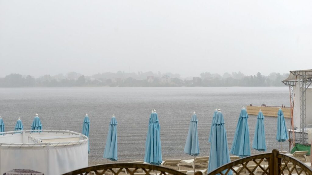 A lakeside resort on a rainy day with closed light blue umbrellas lined up along the shore. The water appears dark and rippled from the raindrops, while the overcast sky and mist obscure the distant landscape. The empty lounge chairs and wet surfaces add to the tranquil yet melancholic atmosphere