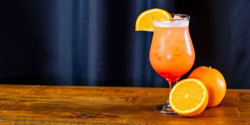 An enticing cocktail with a gradient of orange to red colors is garnished with an orange slice on a wooden countertop, with a dark backdrop enhancing the drink's vibrant colors, suggesting a refreshing citrus beverage.