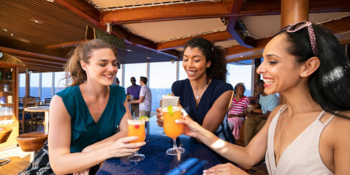 Three women enjoy tropical drinks at a cruise ship bar, with two of them clinking glasses and smiling. The atmosphere is leisurely and social, with other passengers visible in the background, suggesting a vibrant vacation scene on deck.