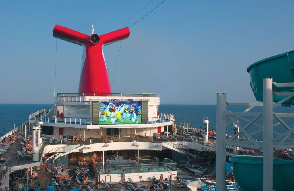 The deck of Carnival Freedom bustling with activity, featuring the signature red funnel and a large outdoor screen displaying a sports event. Passengers relax on sun loungers, some watching the screen, others enjoying the water slide, all against the backdrop of the open sea, exemplifying the leisure and entertainment options available on a cruise vacation.