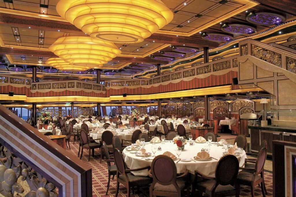 The dining room on Carnival Freedom, featuring a grand and opulent design with large, layered circular light fixtures casting a warm glow over the room. Elegantly set tables are ready to welcome guests, and the richly decorated space boasts intricate patterns and a luxurious ambience, reflecting the ship's commitment to a memorable dining experience.