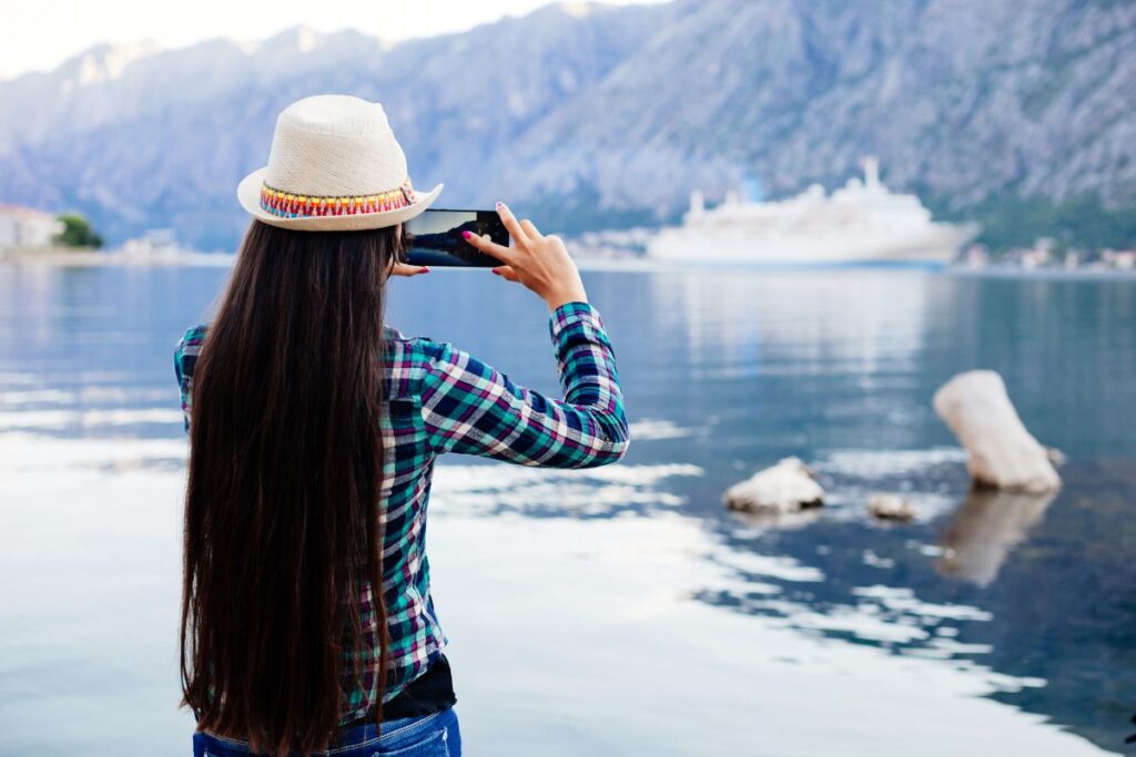 Woman in a plaid shirt and straw hat taking a photo of a mountainous lake scene with a cruise ship in the background, using her smartphone.