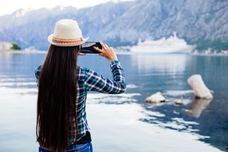 Woman in a plaid shirt and straw hat taking a photo of a mountainous lake scene with a cruise ship in the background, using her smartphone.
