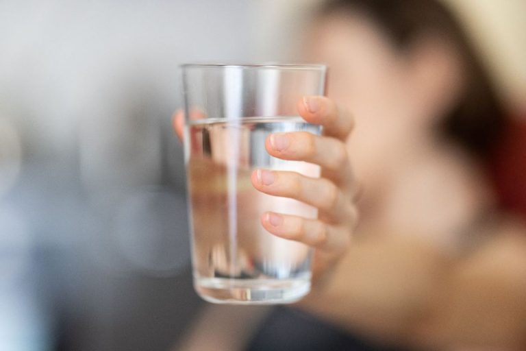 A close-up of a hand holding a clear glass of water, with the focus on the fingers gripping the glass, set against a softly blurred background, highlighting the refreshing simplicity of staying hydrated.