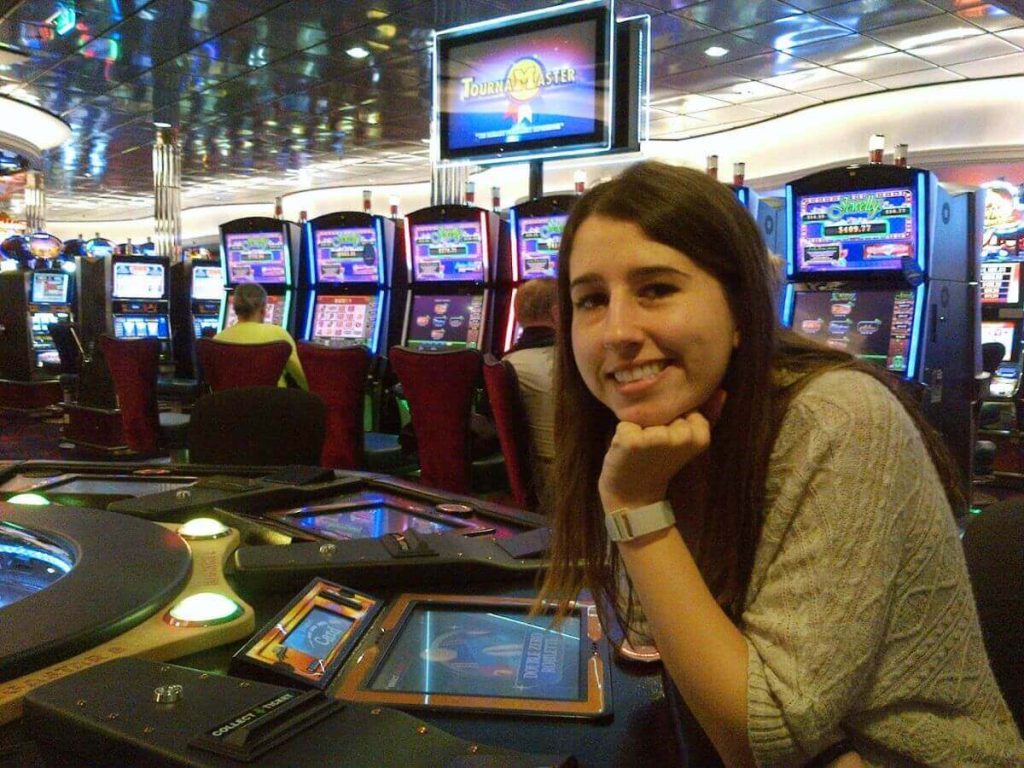 A smiling woman leaning on a gaming table in the vibrant Royal Caribbean casino, surrounded by colorful slot machines and the lively atmosphere of the gaming floor.