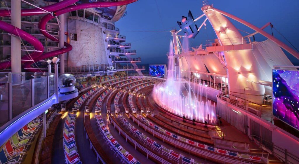 The AquaTheatre on Harmony of the Seas during twilight, featuring a dazzling water show with fountains illuminated by purple lights, surrounded by an outdoor amphitheater with tiered seating and a pink water slide overhead.