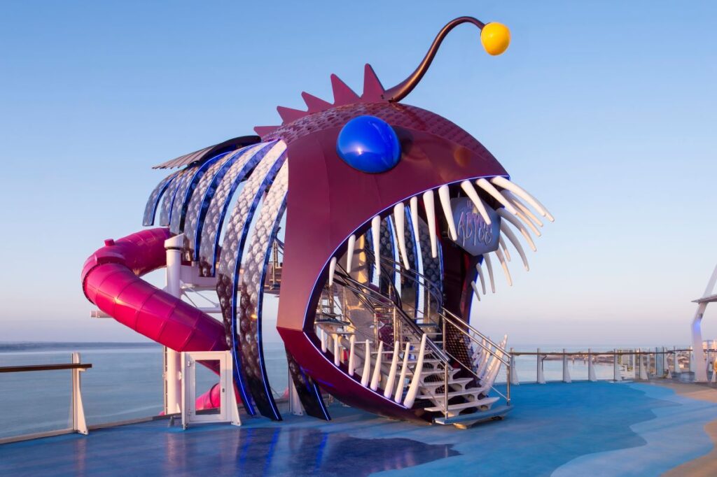 The Ultimate Abyss slide entrance on Royal Caribbean's Harmony of the Seas, depicted as a towering angler fish sculpture with shiny scales and a whimsical design, against a clear sky backdrop with the ocean horizon in the distance.