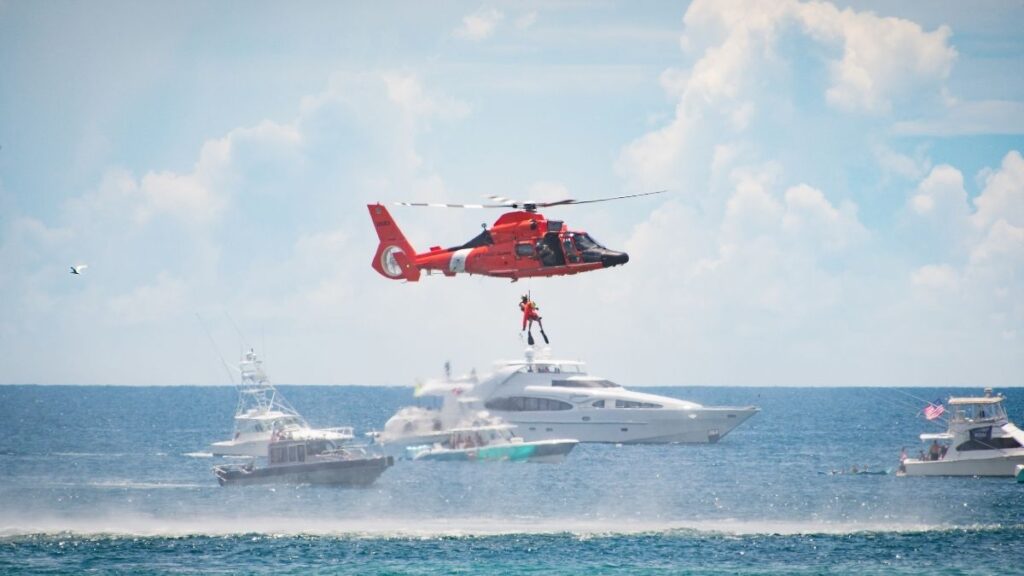 A bright red Coast Guard helicopter hovers above the ocean, lowering a rescue worker on a harness toward the water. Several boats and yachts are in the background, with waves disturbed by the helicopter's rotor wash. The clear blue sky and scattered clouds create a dramatic backdrop for the rescue operation.
