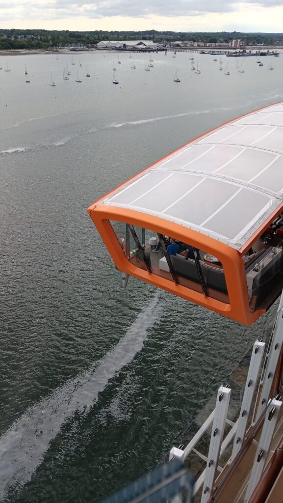 The Celebrity Apex cruise ship's Magic Carpet, an orange, glass-enclosed platform extending over the side of the ship. The Magic Carpet overlooks a calm bay with numerous small sailboats, offering a unique vantage point for passengers.
