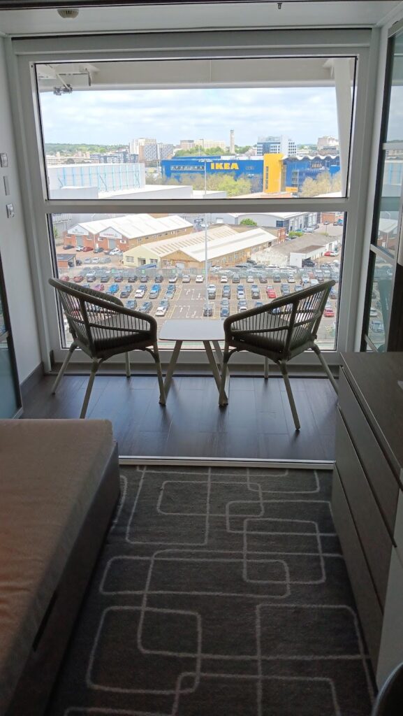 A cozy balcony seating area in a stateroom on the Celebrity Apex cruise ship, offering a view of a cityscape with an IKEA store in the background. The setup includes two chairs and a small table, ideal for enjoying the scenery.