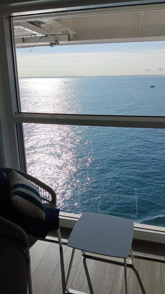 A serene ocean view from a stateroom balcony on the Celebrity Apex cruise ship, with the sun reflecting on the water. The balcony features a chair with a striped pillow and a small table, perfect for relaxing and enjoying the sea.